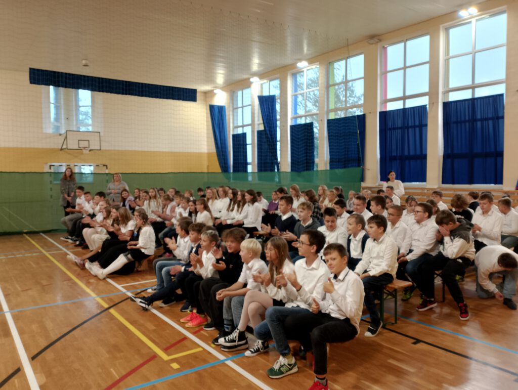 Koncert filharmonii. Na zdjęciu uczniowie klas 4-6 ubrani w galowy strój siedząc na ławeczkach słuchają koncertu filharmonii. 