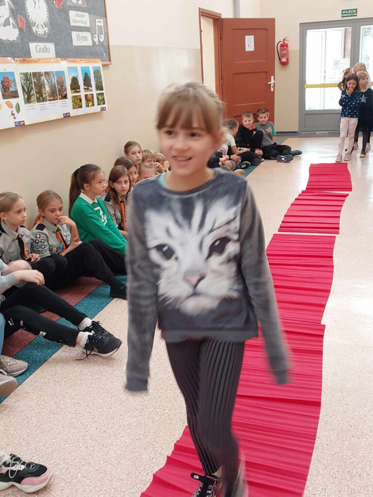 Szkole obchody Międzynarodowego  Dnia Dziewczynek. Na zdjęciu uczennica klasy pierwszej idzie po czerwonym dywanie.  W tle inne dzieci z klas 1-3. 