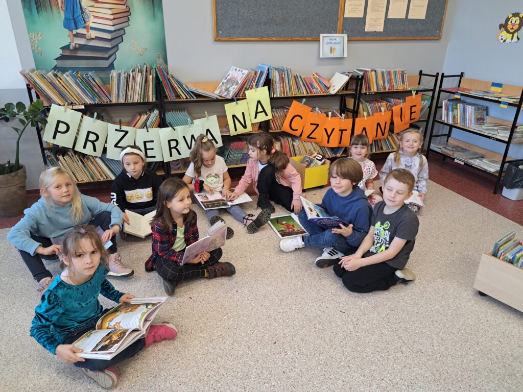 Uczniowie klasy 1a czytają w bibliotece szkolnej wypożyczone przez siebie książki. W tle regały i kolorowy napis Przerwa na Czytanie.