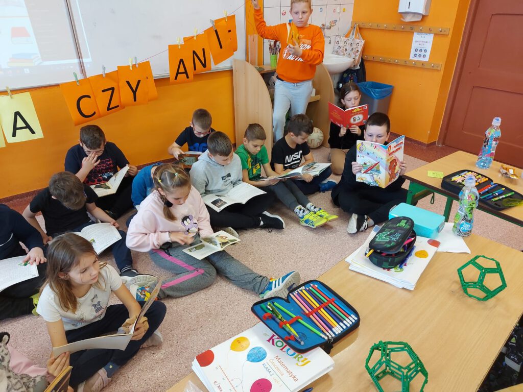 Uczniowie klasy 3a siedzą na podłodze w klasie i wygodnej pozycji czytają książki.