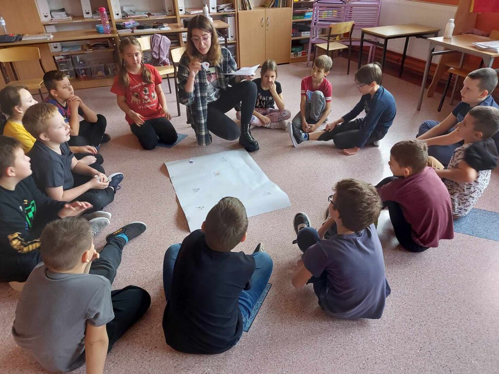Projekt #ActiveSisterSchools. Na zdjęciu nauczycielka tłumaczy uczniom zasady zabawy. Uczniowie siedzą w kołku na podłodze.