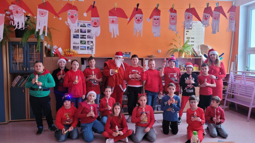 Szkolne mikołajki. Na zdjęciu uczniowie klasy trzeciej ubrani w czerwone stroje i czerwone czapki pozują do zdjęcia z Mikołajem  i z wychowawczynią. 