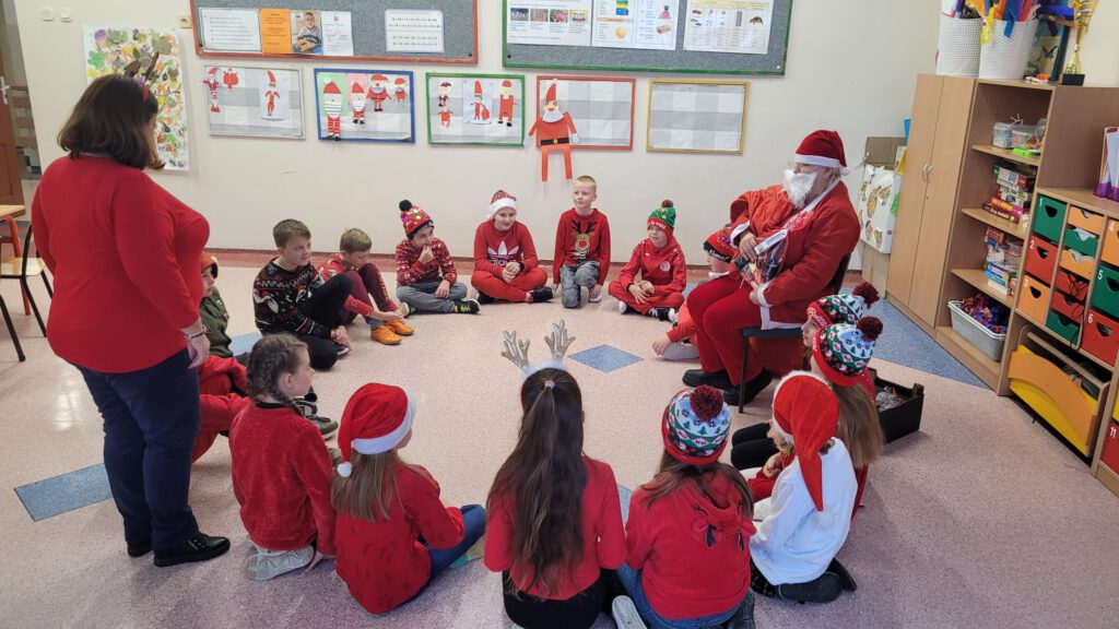 Szkolne mikołajki. Na zdjęciu uczniowie siedzą  w kółku na podłodze, ubrani są  w czerwone stroje i czerwone czapki. Miedzy nimi siedzi Mikołaj.