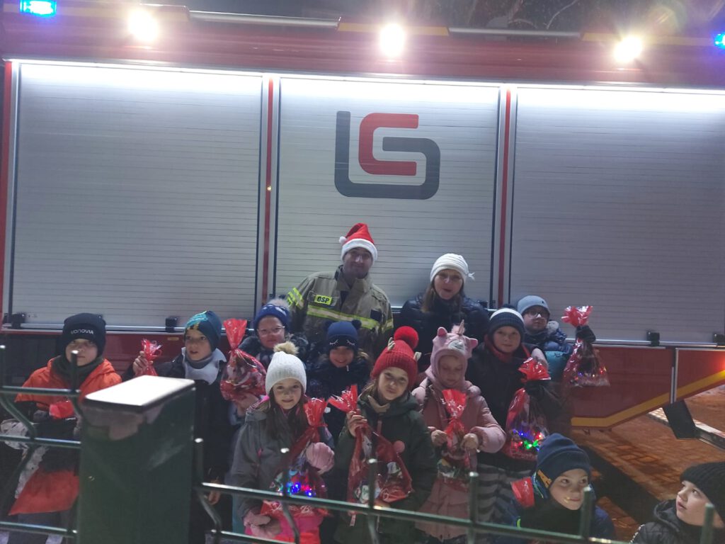 Uśmiechnięte dzieci trzymają w rękach paczki mikołajowe. Za dziećmi stoi wóz strażacki. 