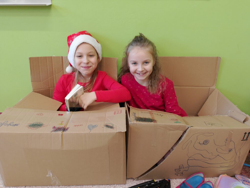 Szkolne mikołajki. Na zdjęciu dwie dziewczynki z klasy pierwszej w czapeczkach czerwonych siedzą w kartonach i je kolorują. 