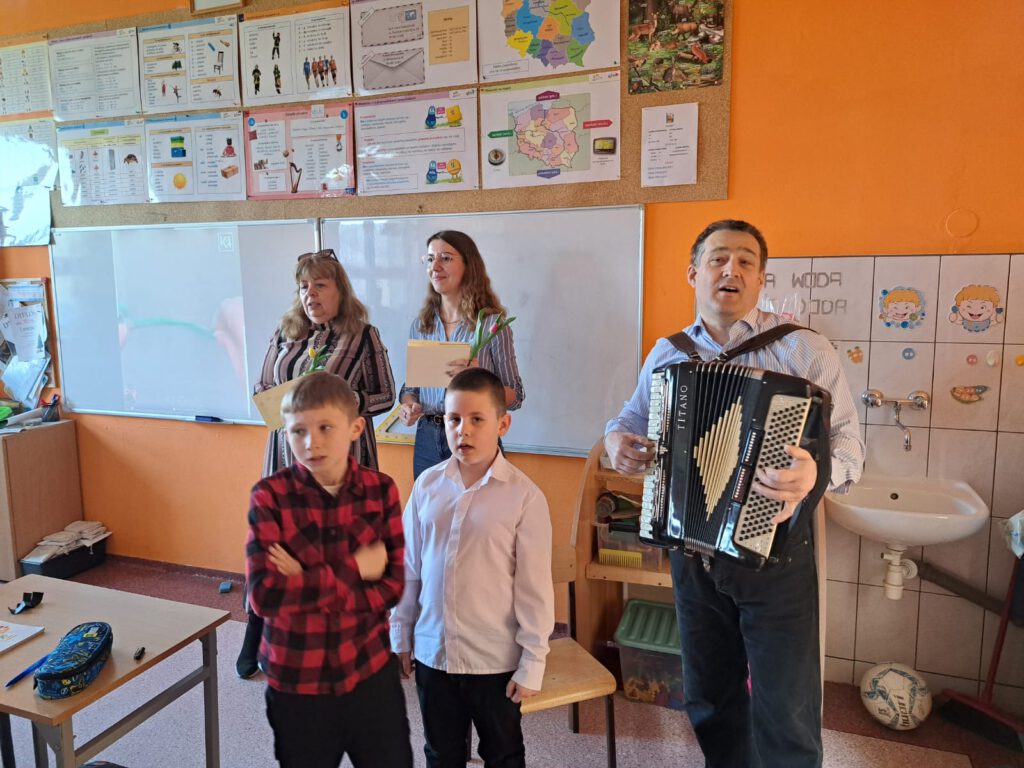 Obchody Dnia  Kobiet w naszej szkole. Na zdjęciu uczniowie składaj życzenia nauczycielce, nauczyciel przygrywa na akordeonie.