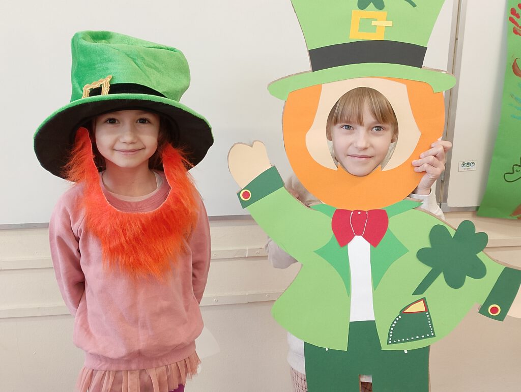 Obchody Dnia św. Patryka. Na zdjęciu dziewczynki pozują do zdjęcia. Jedna z nich ma na głowie zielony kapelusz, druga  trzyma postać skrzata leprechuana. 