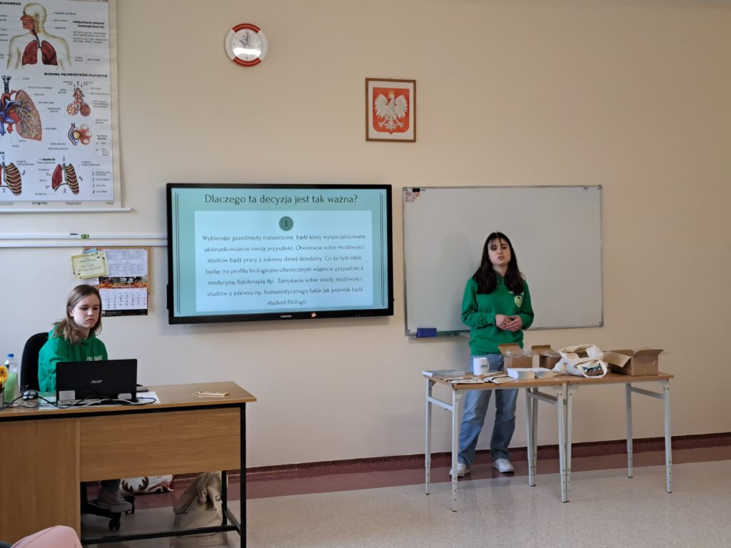 Przedstawia 2 Uczennice w zielonych bluzach z tytułem projektu "Zwolnieni z Teorii ". Jedna z nich stoi przy ławce i mówi o decyzji wyboru zawodu. Druga z nich przełącza slajdy na laptopie.