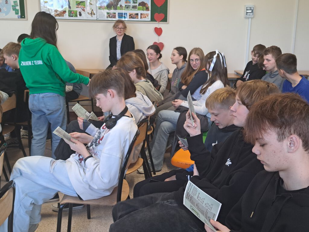 Uczniowie siedzą na krzesłach i czytają z uwagą otrzymane ulotki dot. szkoły ponadpodstawowej.
