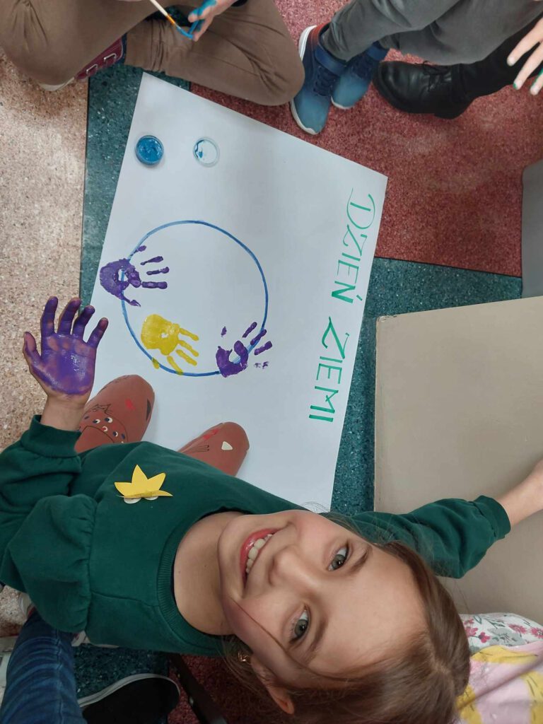 Plakat na Dzień Ziemie "Ziemia w Twoich rękach". Na zdjęciu uśmiechnięta  dziewczynka maluje dłoń farbą. 