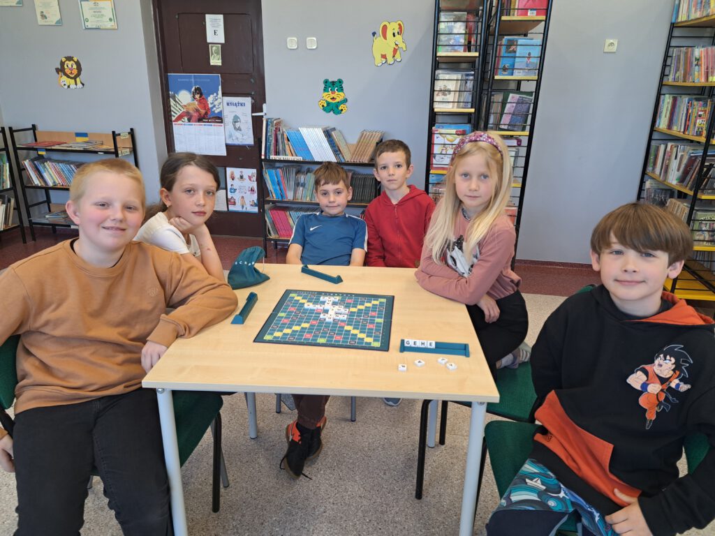 Zdjęcie przedstawia uczniów klas 1-3 podczas zTurnieju Scrabble. Na środku ławki jest plansza do gry Scrabble, obok niej zielony woreczek z literami. Uczniowie mają uśmiechnięte miny.
W tle zdjęcia są regały z książkami. 