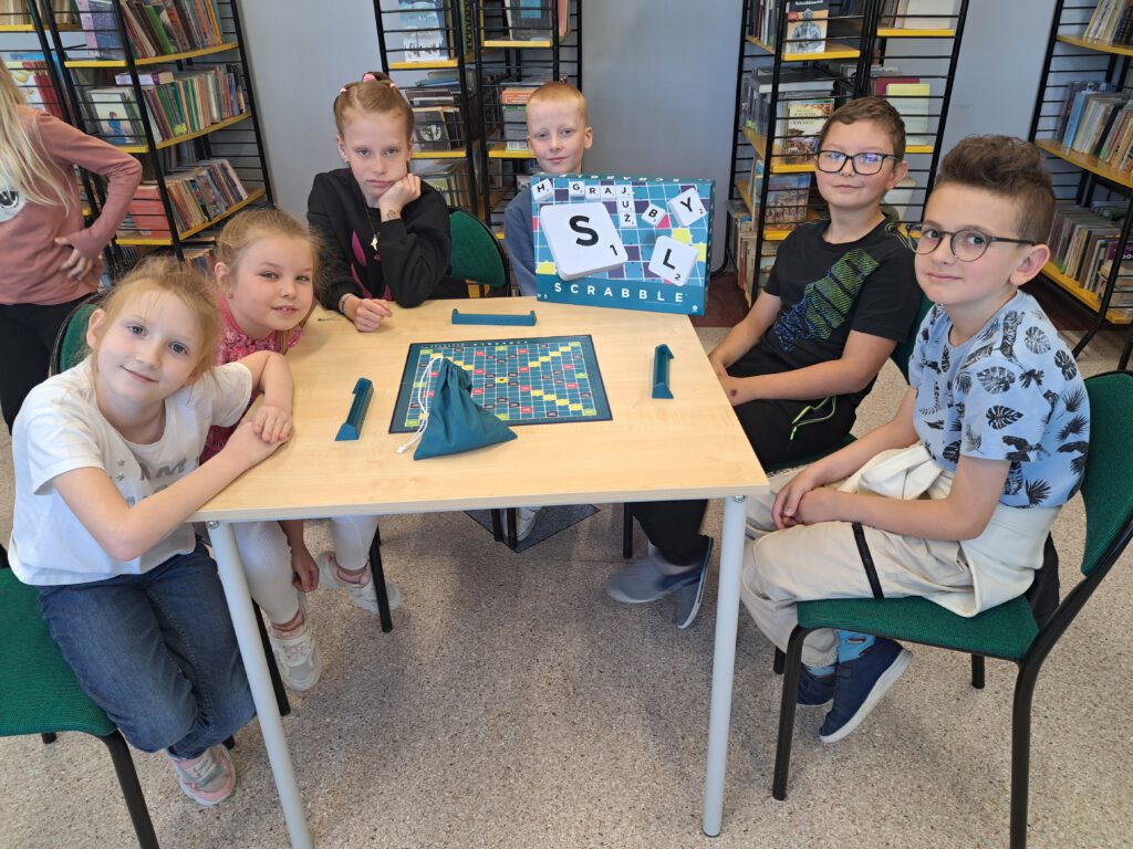 Zdjęcie przedstawia uczniów klas 1-3 podczas zTurnieju Scrabble. Na środku ławki jest plansza do gry Scrabble, obok niej zielony woreczek z literami. Uczniowie mają uśmiechnięte miny.
W tle zdjęcia są regały z książkami. 