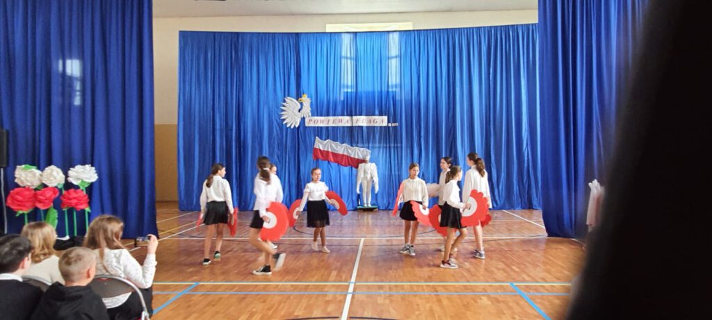 Apel z okazji świat majowych. Na zdjęciu uczennice klasy piątej ubrane w strój galowy i trzymając biało-czerwone pióropusze z papieru wykonują taniec.
