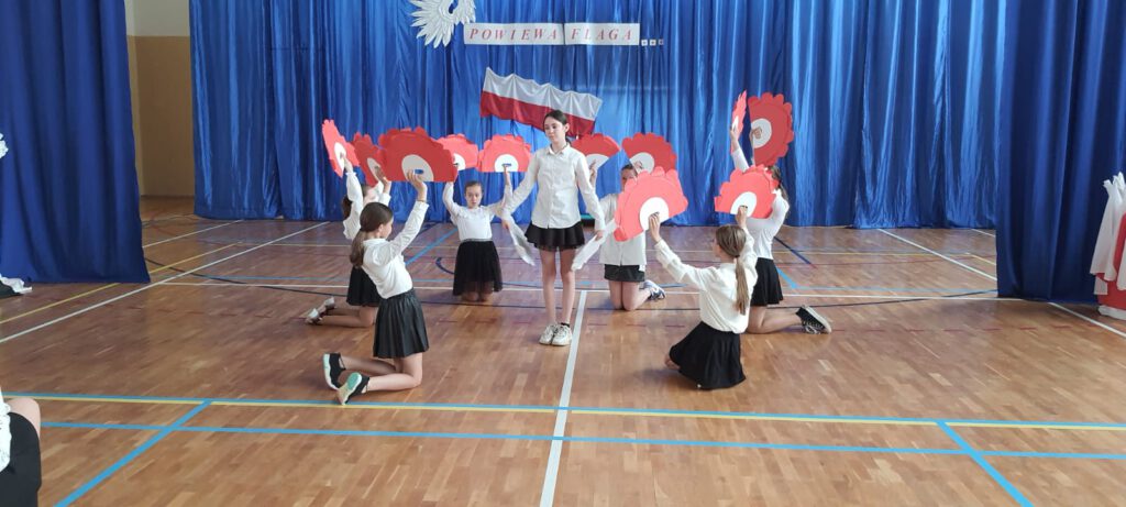 Apel z okazji świat majowych. Na zdjęciu uczennice klasy piątej ubrane w strój galowy i trzymając biało-czerwone pióropusze z papieru wykonują taniec.