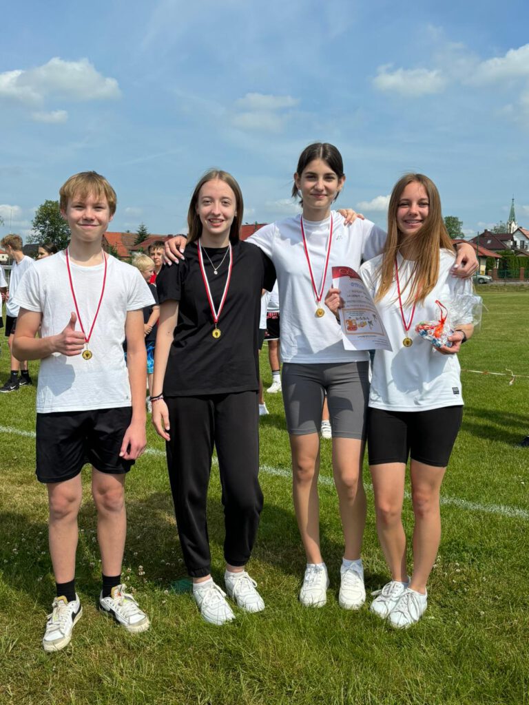 Pietruszkowa sztafeta.  Na zdjęciu uczniowie klasy siódmej - trzy dziewczynki i jeden chłopiec- ubrani w strój sportowy dumnie prezentują zdobyć w biegu medale. 