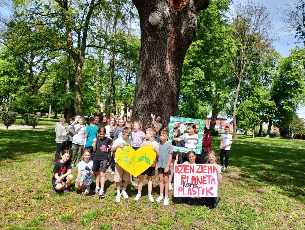 Na zdjęciu uczniowie stoją w parku pod drzewem. dwóch chłopców trzyma żółte serce z napisem " Z miłości do ziemi", obok dwie dziewczynki trzymają plakat z napisem "Dzień Ziemi- planeta kontra plastik", inne dwie uczennice trzymają ramkę z obrazkami na temat   Dnia Ziemi. 