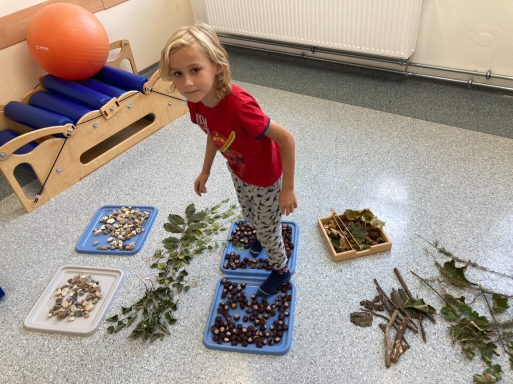 Projekt "Gramy zmysłami"- październik- sensoryczny tor przeszkód. Na zdjęciu uczeń pokonuje tor przeszkód wykonany z różnych materiałów: szyszek, żołędzi, liści, kamieni. 