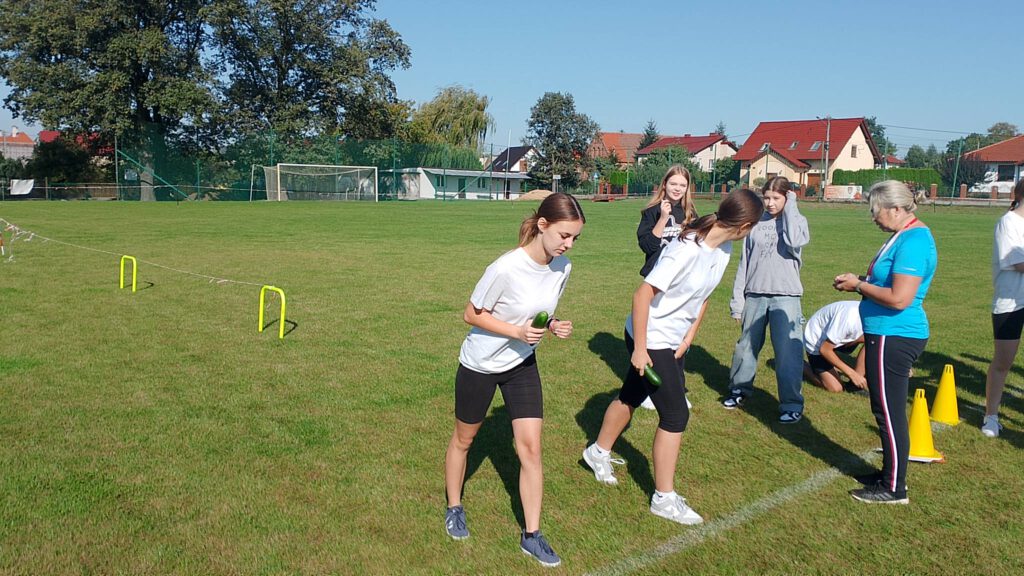 Ogórkowa sztafeta- szkolna akcja sportowa. Na zdjęciu dwie uczennice przygotowują się do biegu. Nauczycielka wychowanie fizycznego przygotowuje stoper, aby zacząć odmierzać czas. 