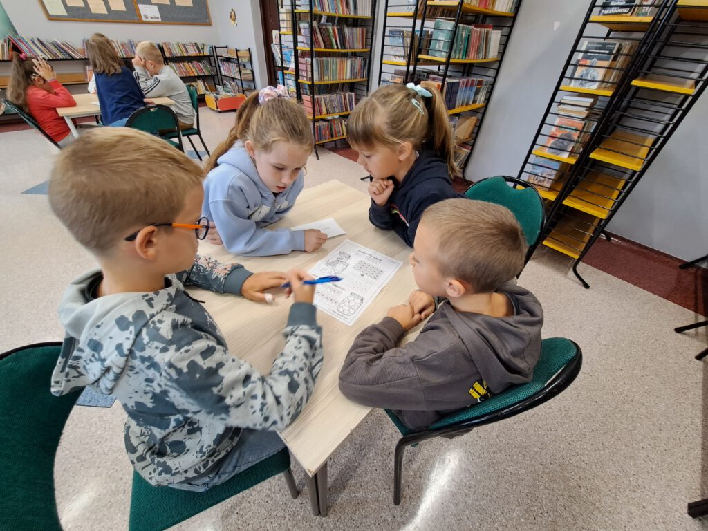 Przedstawia uczniów siedzących przy ławce. Rozwiązują oni łamigłówki.  W tle zdjęcia inna grupa dzieci oraz regały z książkami. 