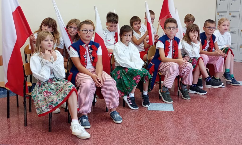 Projekt #Active Sister Schools. Na zdjęciu uczniowie klasy trzeciej b ubrani w biało-czerwone stroje trzymając biało-czerwone flagi  czekają na losowanie drużyn w projekcie. 