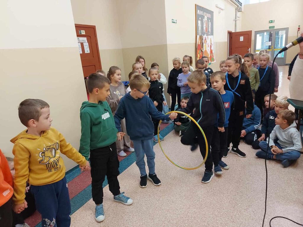 Uczniowie z klasy 1a biorący udział w konkurencji polegającej na przeprowadzeniu hula hopu z jednego końca szeregu na drugi. Uczniowie trzymają się za ręce i po kolei przechodzą przez obręcz.