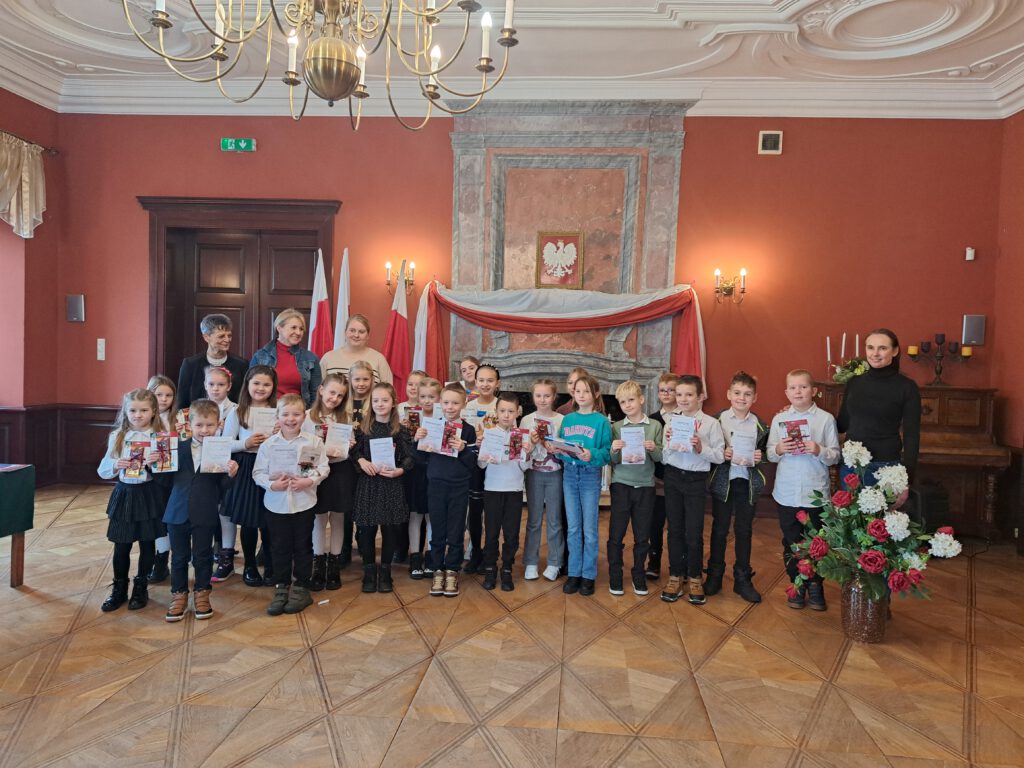 Przedstawia uczestników etapu gminnego Recytatorskiego Konkursu Patriotycznego "Młode Pokolenie".
Uczniowie trzymają w rękach dyplomy oraz nagrody.  Obok dzieci stoją członkowie jury i pani dyrektor GOKiB w Gaworzycach. W tle kominek, godło oraz przywieszona flaga biało-czerwona. Obok dzieci znajduje się wazon z biało-czerwonymi kwiatami. 
