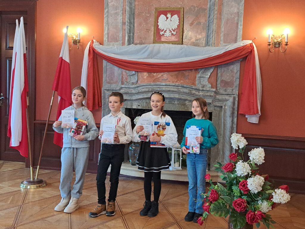 Przedstawia laureatów etapu gminnego Recytatorskiego Konkursu Patriotycznego "Młode Pokolenie".
Uczniowie trzymają w rękach dyplomy oraz nagrody.  W tle kominek, godło oraz przywieszona flaga biało-czerwona. Obok dzieci znajduje się wazon z biało-czerwonymi kwiatami. 