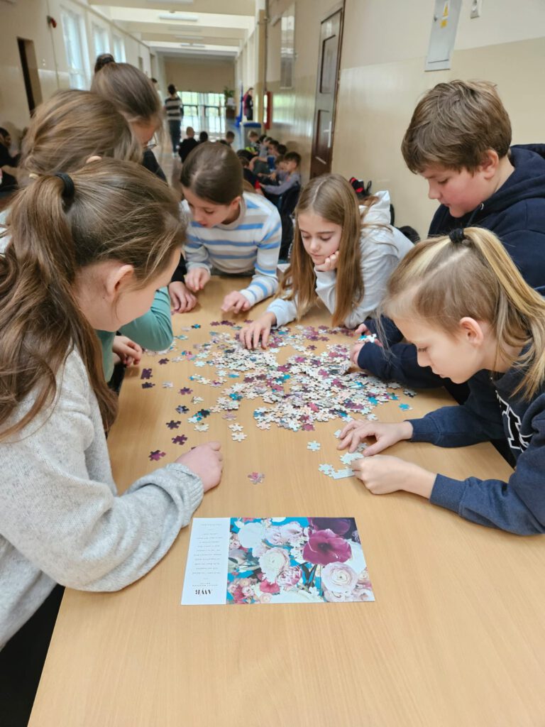 Na zdjęciu grupa uczniów zgromadzonych wokół dużego stołu.  Układają wspólnie puzzle. 