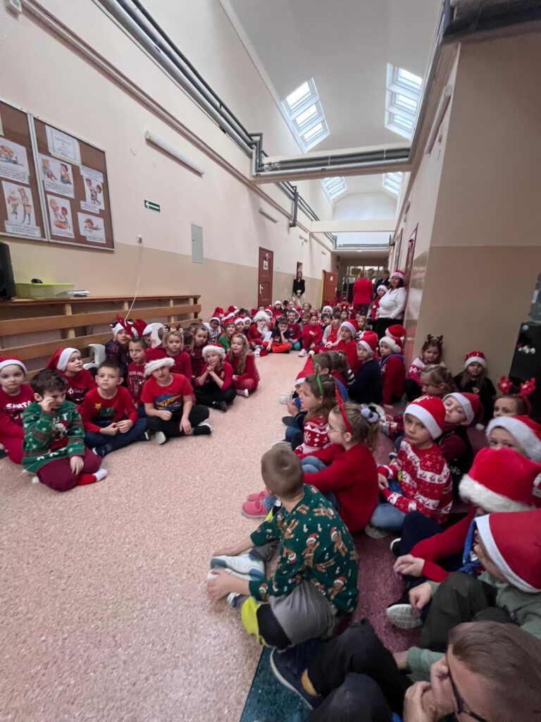 Koncert piosenek świątecznych w języku angielskim. Na zdjęciu uczniowie edukacji wczesnoszkolnej ubrani w czerwone świąteczne stroje z czapkami  mikołajowymi na głowie siedzą na korytarzu i czekają na swój występ.  