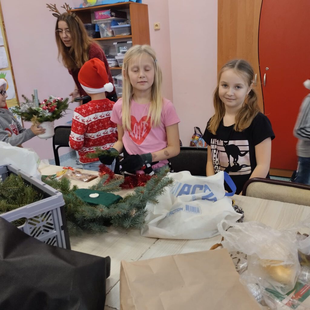 Przygotowania do świąt- stroiki świąteczne. Dziewczynki z klasy 2a wykonują stroik. Jedna z nich ma założone rękawiczki. W tle wychowawca świetlicy pomaga młodszym dzieciom.