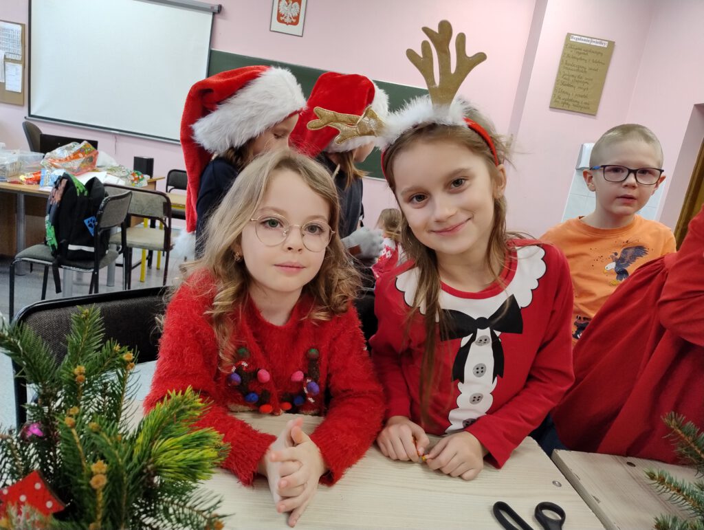 Przygotowania do świąt- stroiki świąteczne. Dwie dziewczynki z klasy 1a pozują do zdjęcia. Mają na sobie czerwone stroje, jedna z nich ma założone rogi renifera.