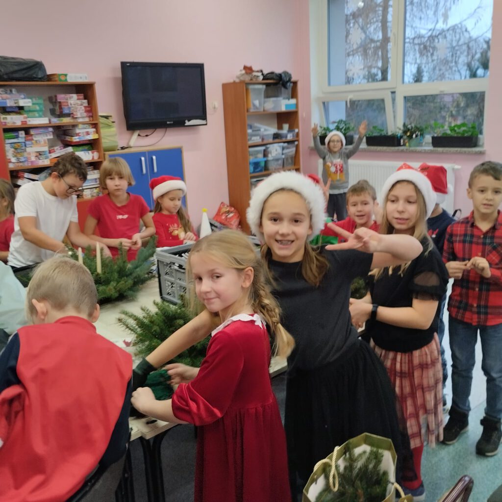 Przygotowania do świąt- stroiki świąteczne. Uczniowie w czerwonych strojach i czapkach Mikołaja stoją przy złączonych stołach i pracują nad stroikami.