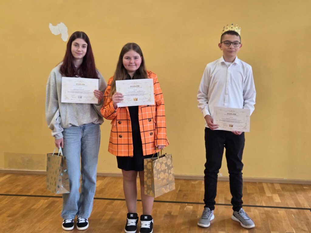 2 dziewczyny i 1 chłopak, którzy zostali laureatami konkursu Nowy banknot z Bolesławem Chrobrym. Uczniowie w rękach trzymają dyplomy i nagrody w torebkach prezentowych. W tle zdjęcia żółta ściana sali gimnastycznej.