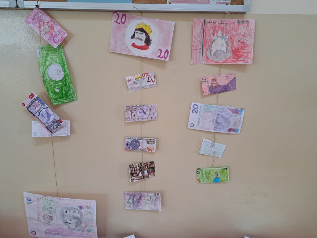 Przedstawia kolorowe banknoty wykonane przez uczniów wiszące na sznurku. Prace są w kolorach: szarym, żółtym, różowym i fioletowym. Prace wiszą na tle żółtej ściany.