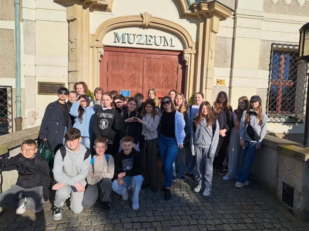 Przedstawia uczestników wycieczki przed wejściem do Muzeum Archeologiczno- Historycznego w Głogowie. W tle zdjęcia stare, brązowe drzwi z napisem Muzeum oraz zakratowane okna budynku.
