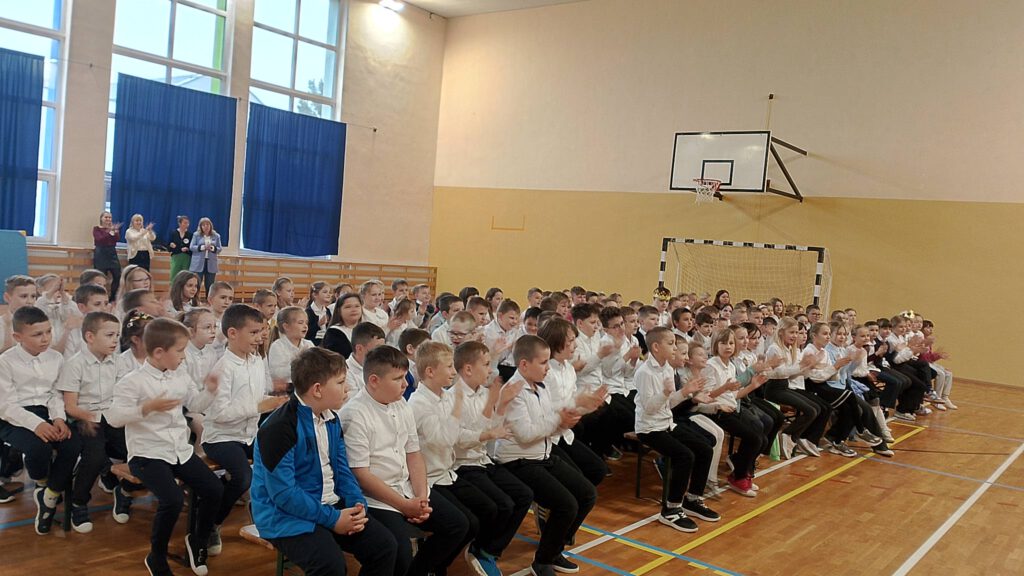 Na zdjęciu uczniowie klas 1-3 ubrani w strój galowy siedzą na ławeczkach w sali gimnastycznej słuchają utworów muzycznych i klaszczą w dłonie.