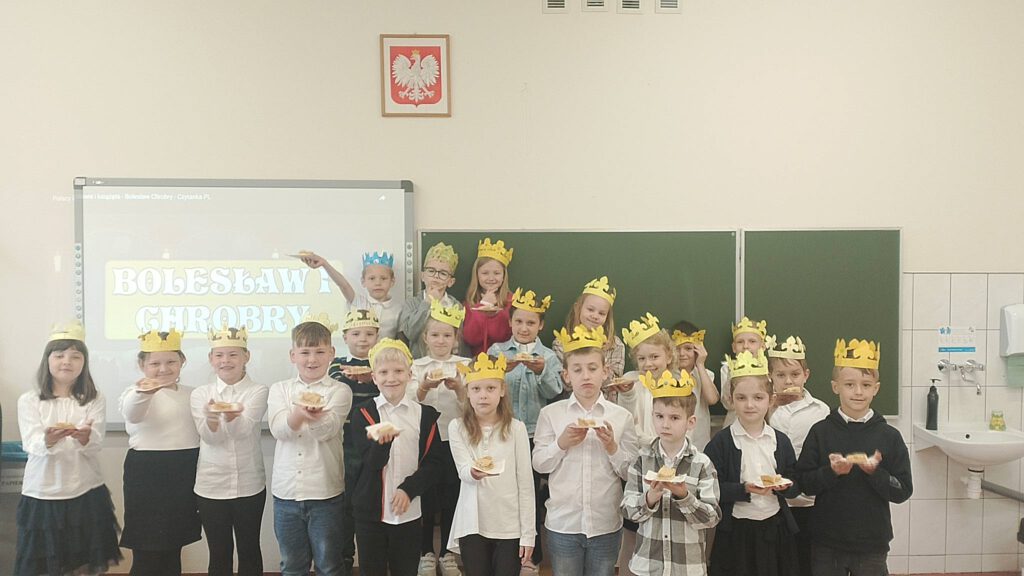 Tysiąclecie koronacji Bolesława I Chrobrego. Na zdjęciu uczniowie klasy pierwszej z galowych strojach pozują do zdjęcia koronami na głowie, w tle tablica napisem Bolesław I Chrobry.