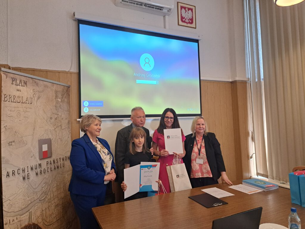 Przedstawia moment wręczenia nagrody i dyplomu uczennicy oraz nauczycielce-opiekunowi konkursu. Przedstawiciele archiwum i Kuratorium Oświaty są uśmiechnięci, stoją wokół uczennicy i nauczycielki. W tle tablica z napisem Archiwum Państwowe we Wrocławiu. Przed osobami na zdjęciu znajduje się biurko na którym położone są dyplomy i nagrody.