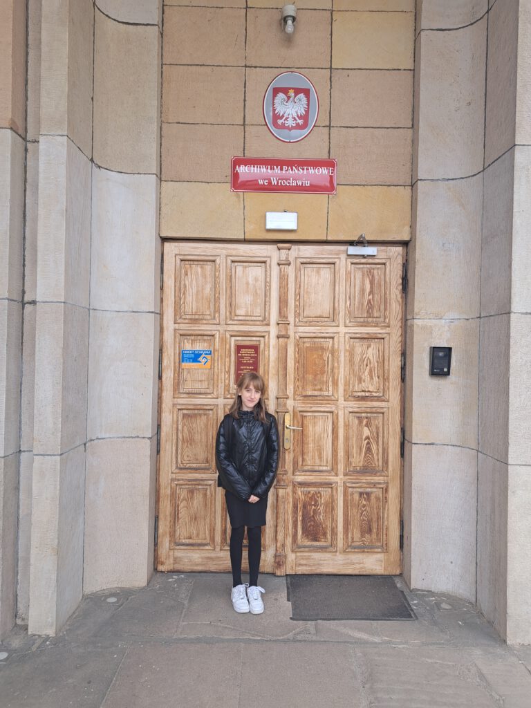 Konkurs "Bez korzeni nie zakwitniesz". Zdjęcie przedstawia Elizę- finalistkę konkursu, ubraną w czarną kurtkę i sukienkę na tle drewnianych drewnianych drzwi głównego wejścia do archiwum.