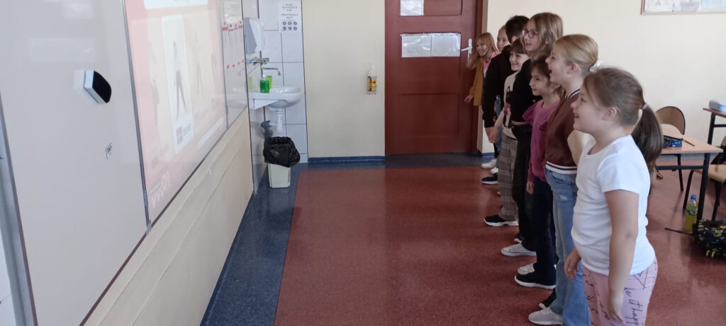 Projekt Active Sister Schools. Na zdjęciu uczniowie klasy trzeciej stojak w jednym rzędzie i wykonują ćwiczenia, które wyświetlają się na tablicy.