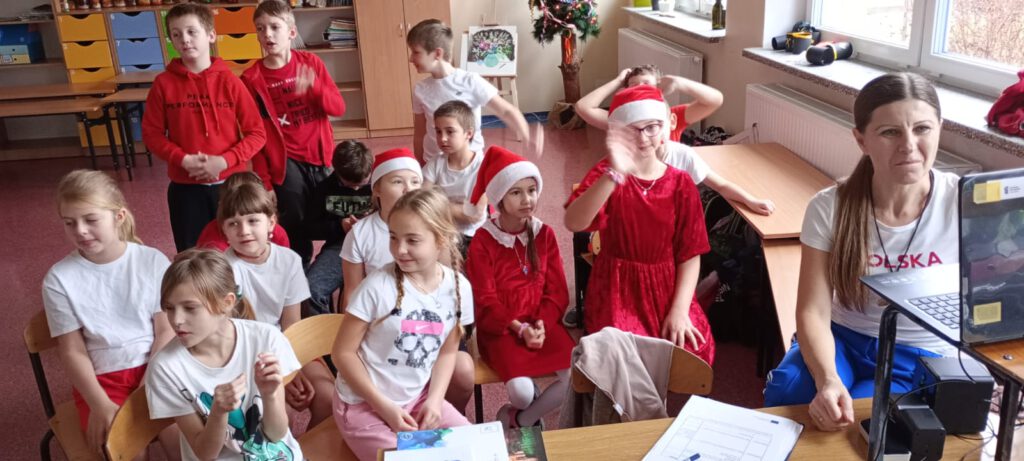Projekt Active Sister Schools. Na zdjęciu uczniowie klasy trzeciej siedzą na podłodze i patrzą na ekran komputera. Część dzieci ubrana jst w strój mikołajkowy.