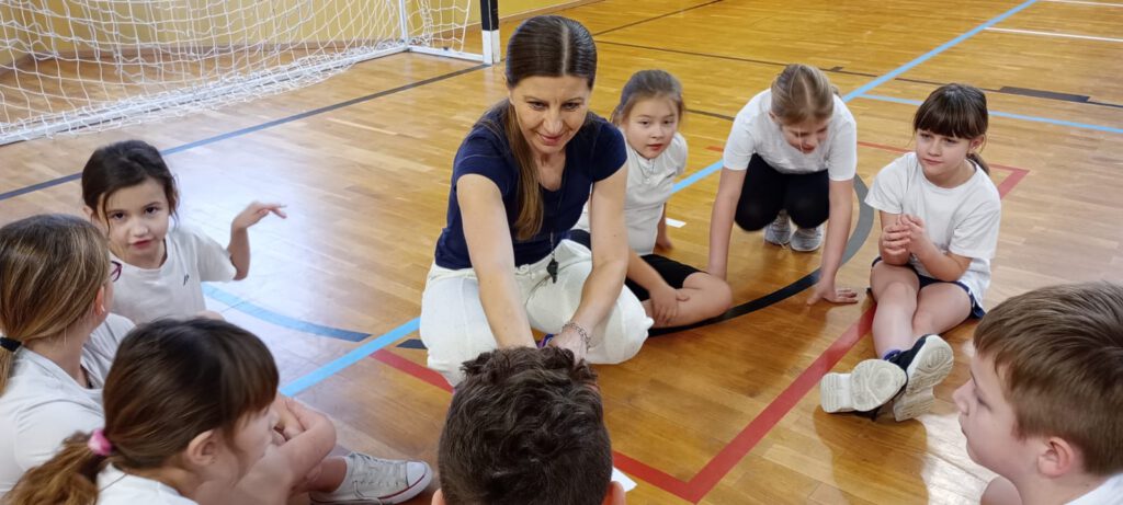 Projekt Active Sister Schools. na zdjęciu uczniowie kasy trzeciej ubrani w strój sportowy siedzą na podłodze na sali gimnastycznej i i słuchają poleceń nauczyciela.