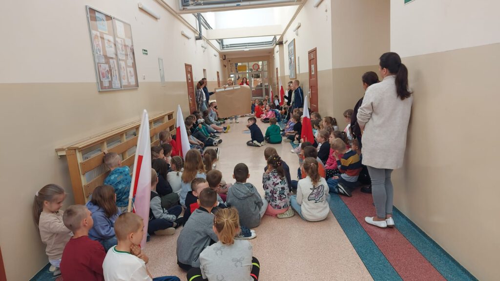 Święta Majowe. Uczniowie klas 1-3 siedzą na korytarzu szkolnym i słuchają przebiegi zajęć związanych ze świętami majowymi.