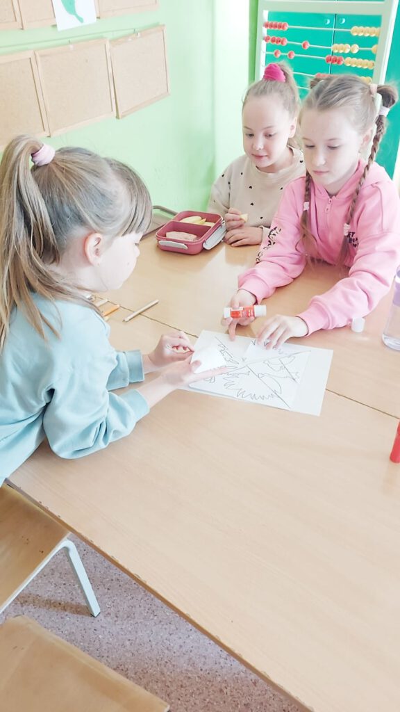 Święta Majowe. Uczennice klasy drugiej siedzą przy ławce szkolnej i rozwiązuję przydzielone im zadania: układają z wyciętych elementów orła.