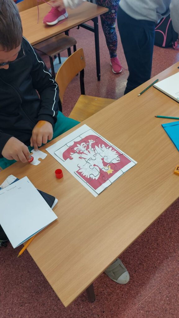 Święta Majowe. Uczeń klasy trzeciej siedzi przy ławce szkolnej i układa godło Polski z puzzli.