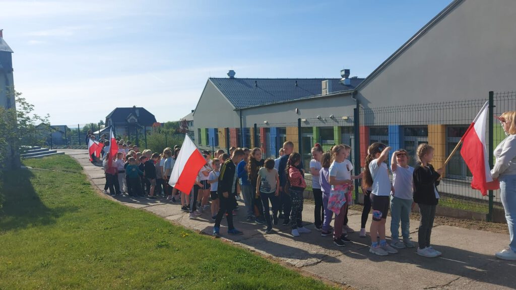Święta Majowe. Uczniowie klas 1-3 trzymając biało-czerwone flagi biorą udział w pochodzie z flagami.