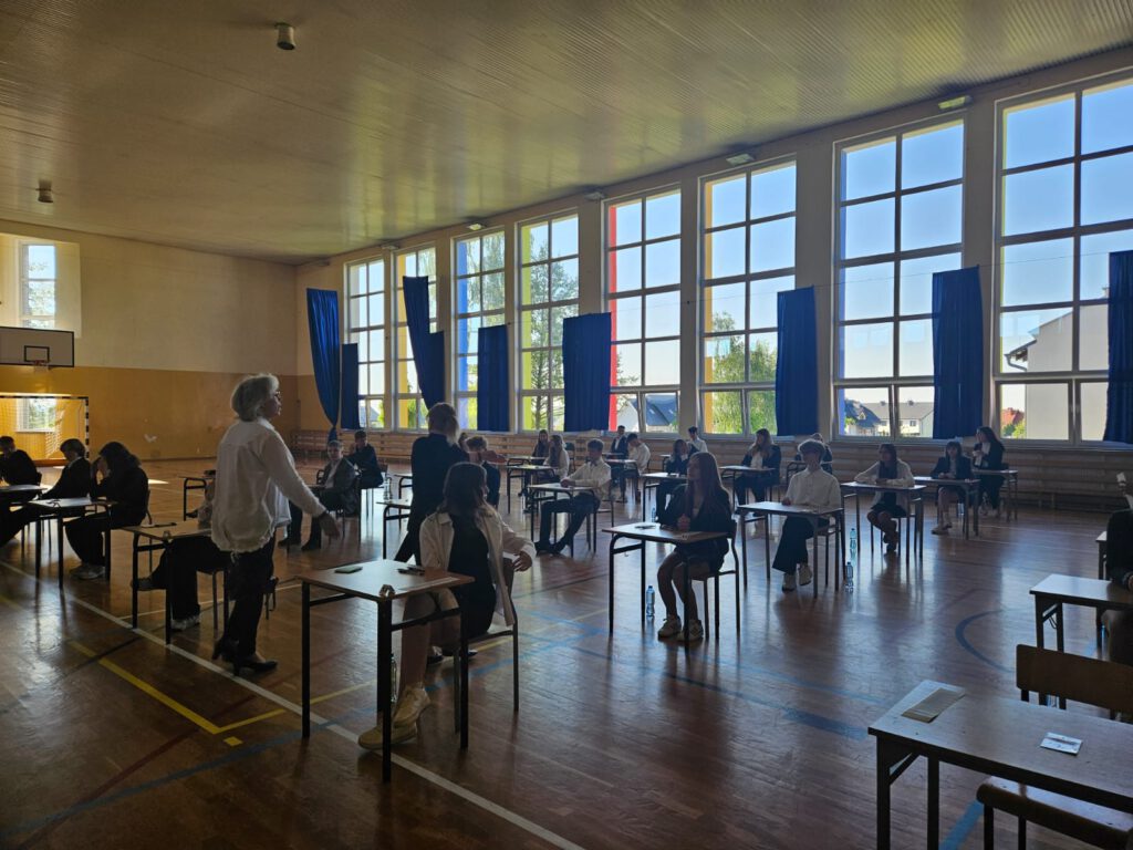Egzamin ósmoklasisty. Na zdjęciu uczniowie klas ósmych ubrani w strój galowy siedzą w ławeczkach na sali gimnastycznej i czekają na rozpoczęcie egzaminu.
