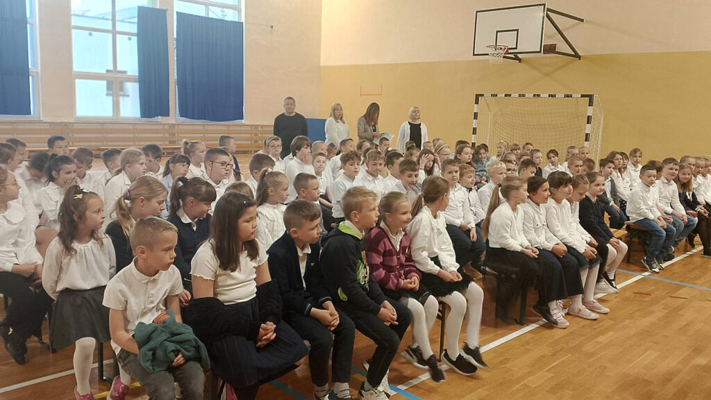 Koncert filharmonii. Na zdjęciu uczniowie klas 1-3 ubrani w strój galowy siedzą na ławeczkach na sali i słuchają koncertu filharmonii.