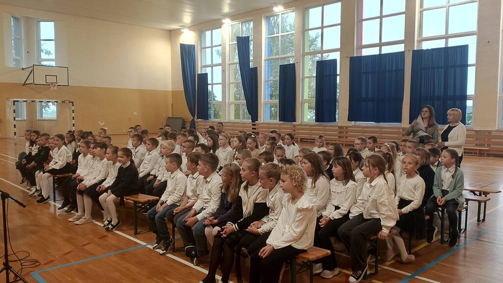 Koncert filharmonii. Na zdjęciu uczniowie klas 1-3 ubrani w strój galowy siedzą na ławeczkach na sali i słuchają koncertu filharmonii.