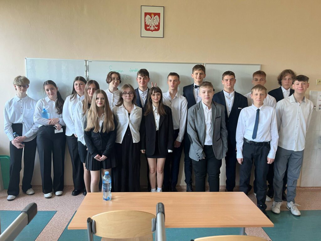 Egzamin ósmoklasisty. Na zdjęciu uczniowie klasy 8 a ubrani na galowo stoją przy ścianie w klasie i pozują do zdjęcia.